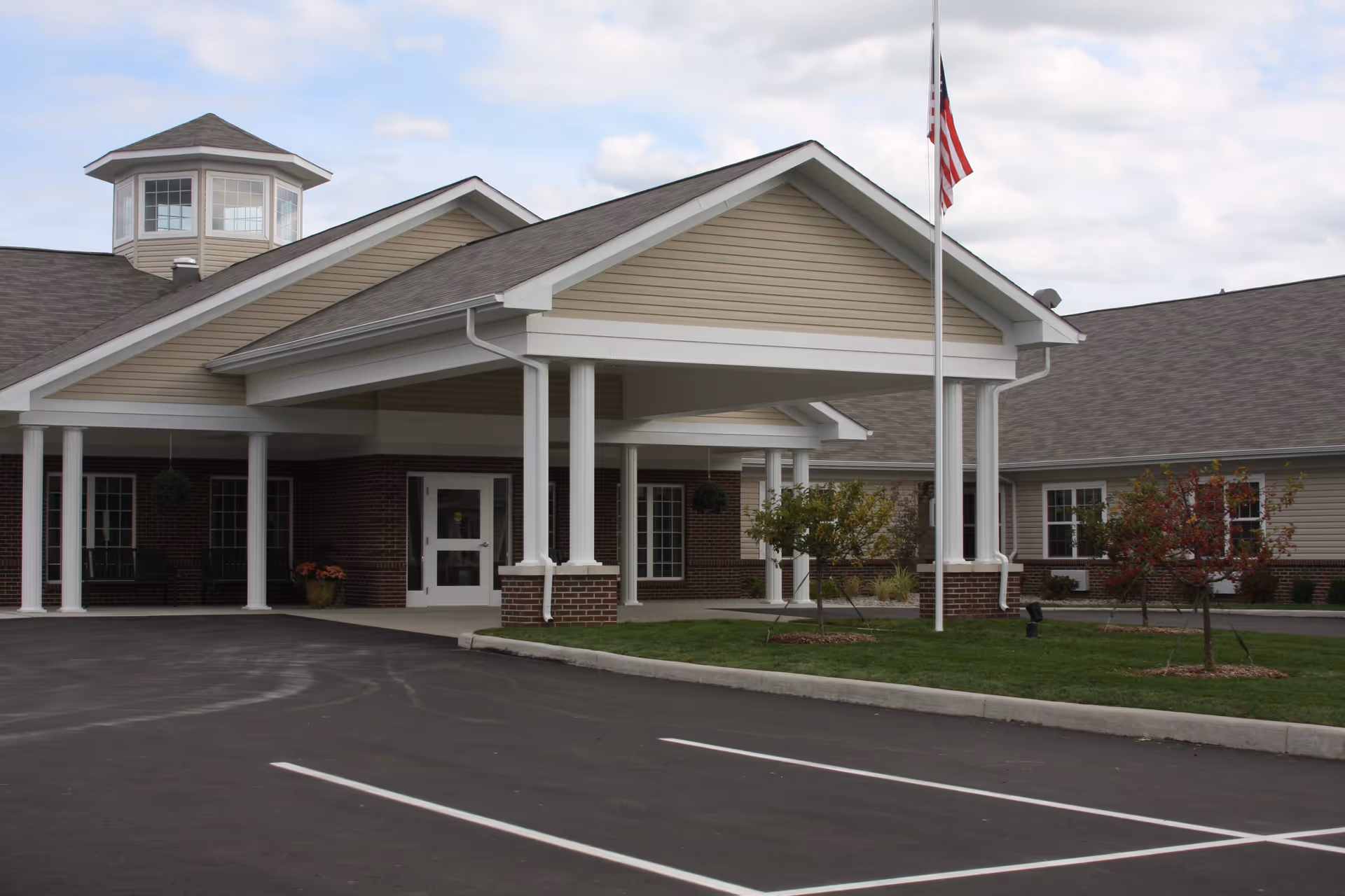 Exterior view of The Oaks at Woodfield building entrance with a covered drop-off area supported by white columns, an American flag on a flagpole, small trees, and a parking lot in front.