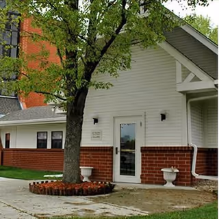 Front entrance of a white-sided, red-brick building with a tree, planters, and a glass entry door.