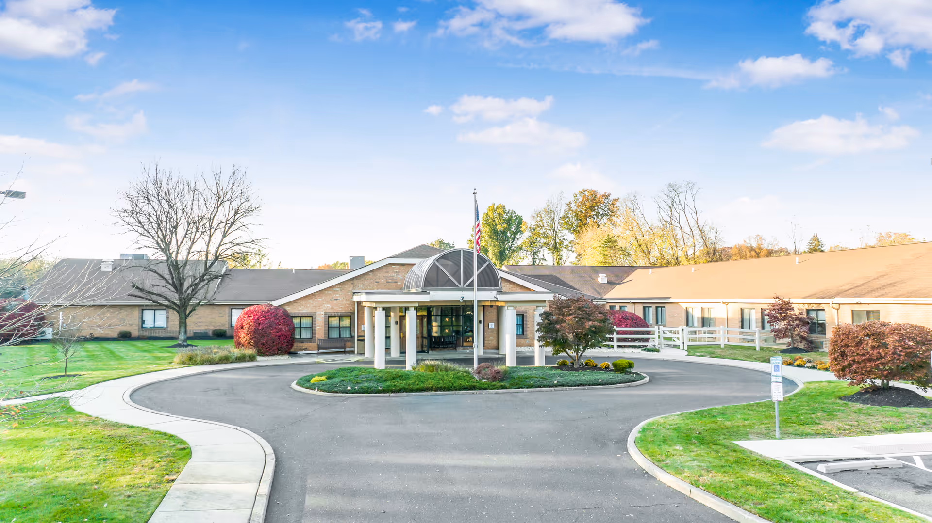 Front exterior view of Yardley Rehabilitation and Healthcare Center showing a single-story building with a covered entrance, a circular driveway, manicured lawns, trees, and an American flag on a flagpole.