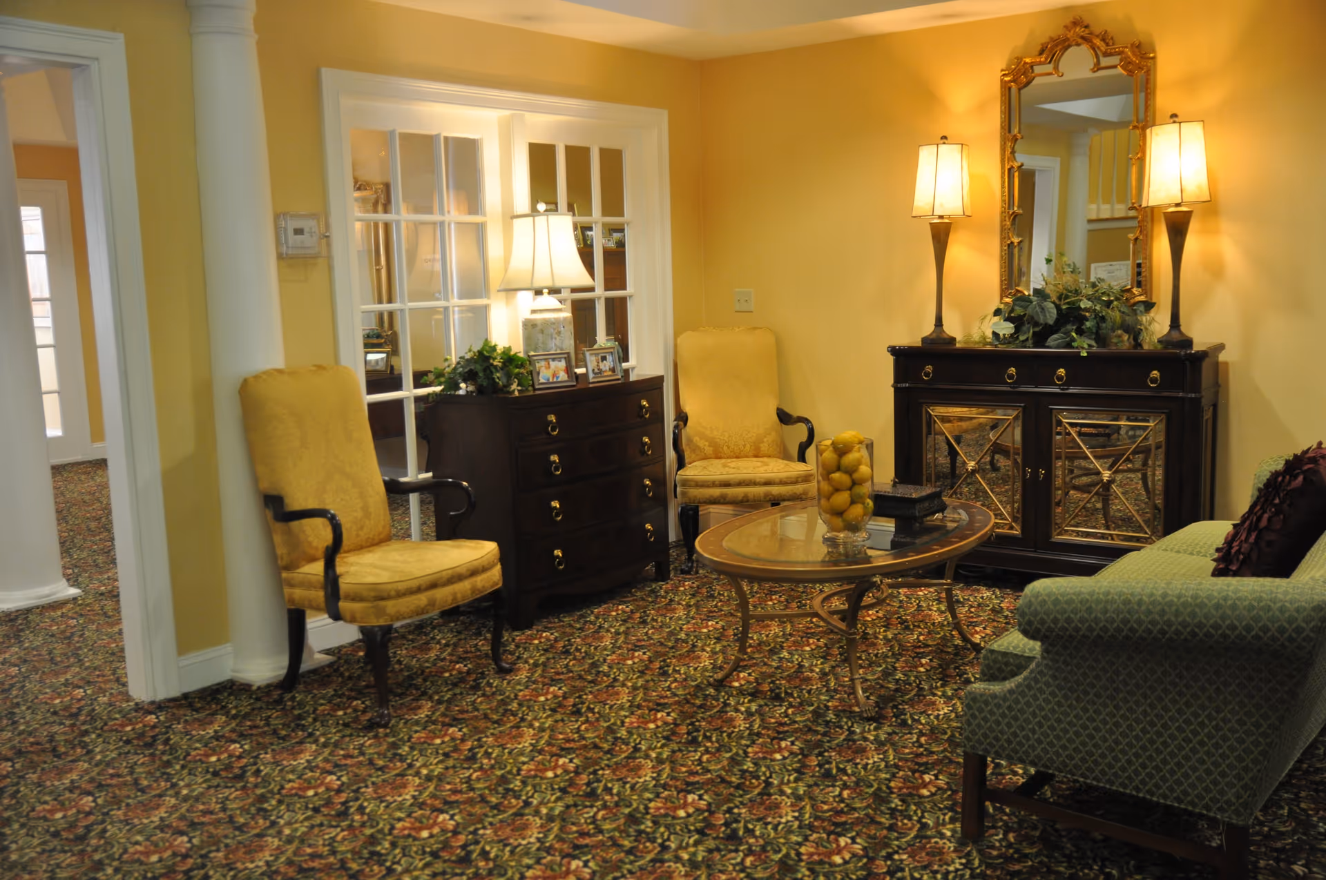 A cozy living room area with two yellow upholstered armchairs, a green patterned sofa with a purple pillow, a glass-top coffee table with a vase of lemons, a dark wooden dresser with a lamp and framed photos, and a matching dark wooden cabinet with a large ornate gold-framed mirror and two lamps on top. The walls are painted yellow and the floor is covered with a floral patterned carpet.