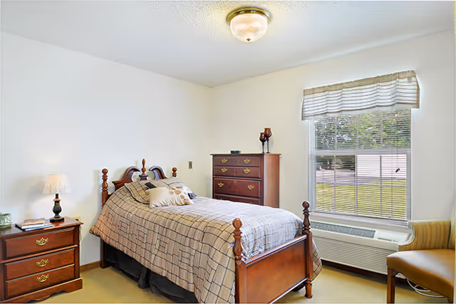 A neatly arranged bedroom with a single wooden bed featuring a checkered bedspread and decorative pillows. Next to the bed is a wooden nightstand with a lamp and some items on it. Across from the bed is a wooden chest of drawers with decorative items on top. A large window with blinds and a valance lets in natural light, and beneath it is an air conditioning unit. The walls are light-colored and the carpet is beige.