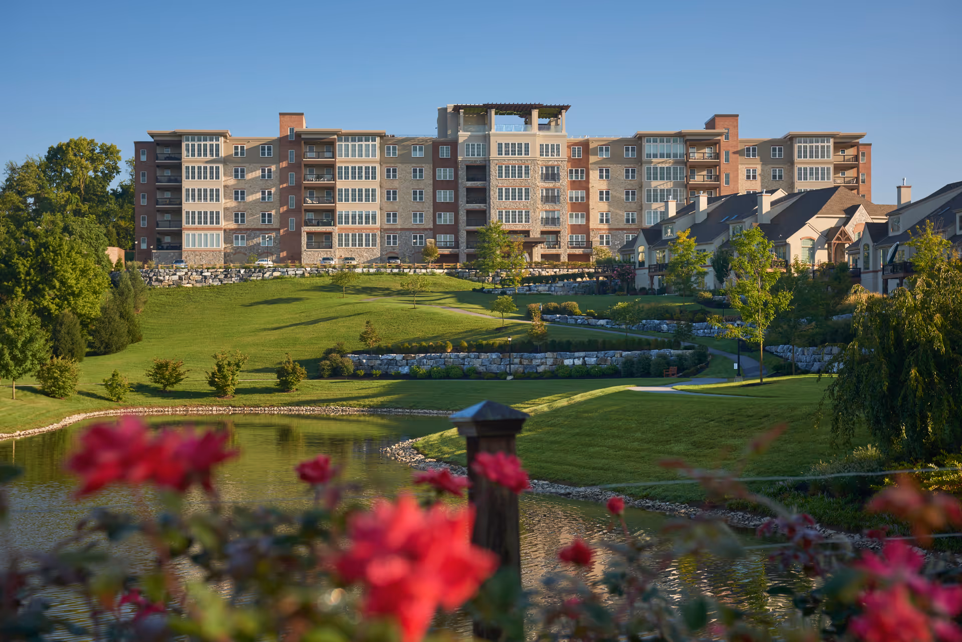 A large multi-story senior living building overlooks landscaped lawns, stone terraces and a pond with red flowers in the foreground.