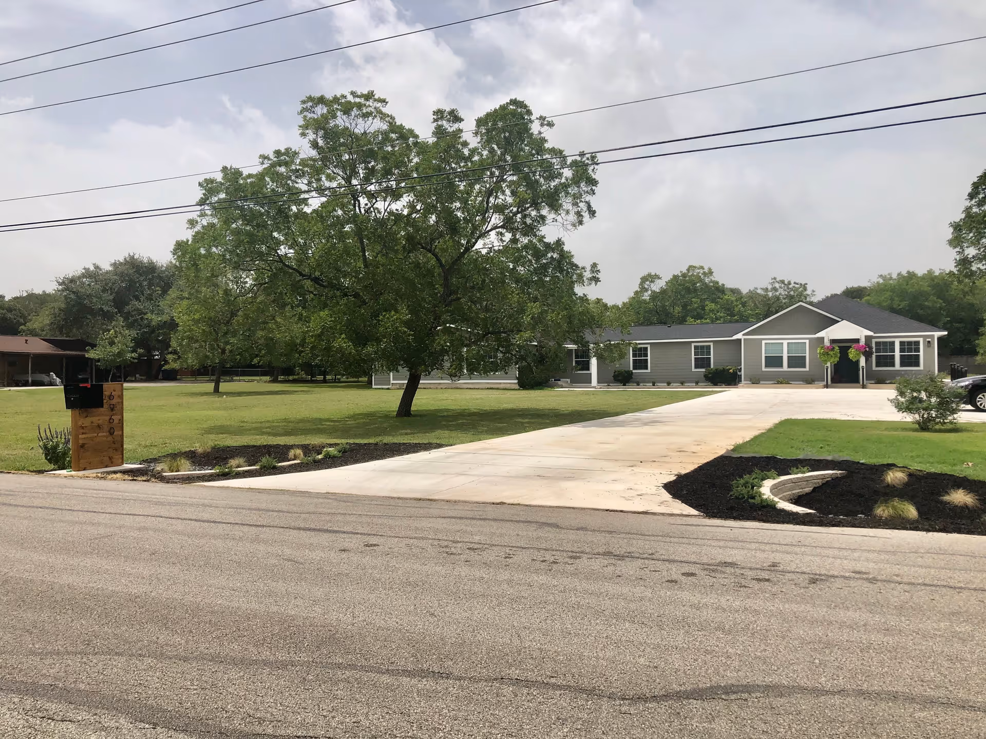 A single-story gray building with white trim, surrounded by a large green lawn and trees. A concrete driveway leads from the street to the building's entrance, which has two hanging flower baskets. There is a wooden mailbox post with the number 69200 near the street.