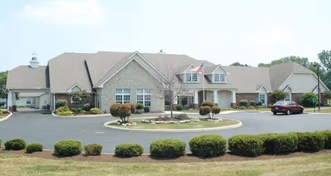 Front exterior view of a single-story senior living facility building with a circular driveway, landscaped bushes, and an American flag near the entrance under a clear sky.