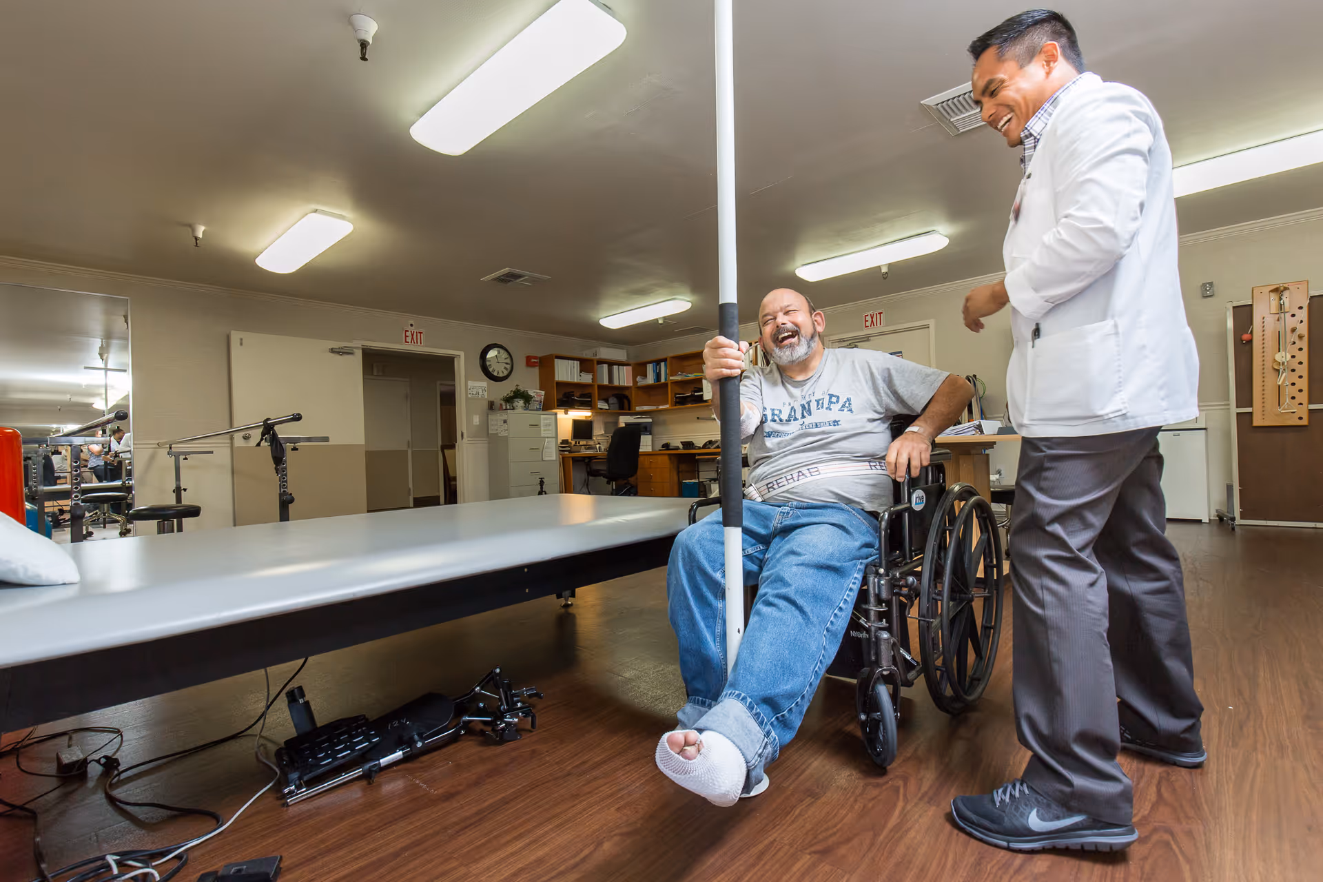 A man in a wheelchair with a bandaged foot is holding onto a pole and laughing while interacting with a healthcare professional in a white coat in a rehabilitation or therapy room with exercise equipment and office furniture.