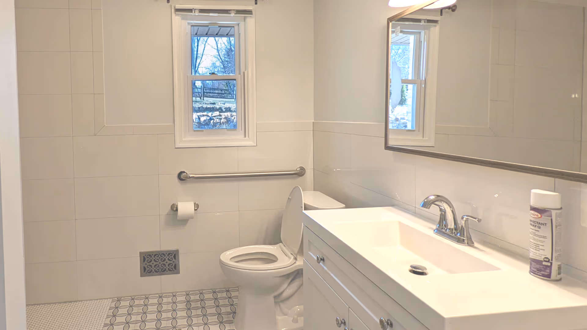 A clean, well-lit bathroom featuring a white toilet with the lid open, a white vanity with a sink and chrome faucet, a large mirror above the sink, a window with a view outside, a wall-mounted grab bar, and a roll of toilet paper on a holder. The floor has patterned tiles and the walls are tiled in white.