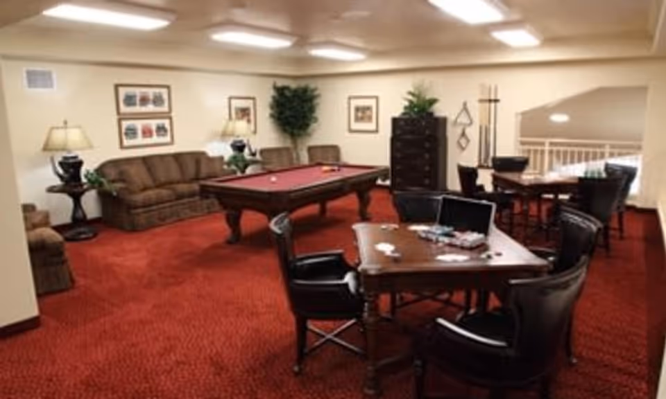 Community game room with a pool table, card tables, sofas and chairs on red carpet.