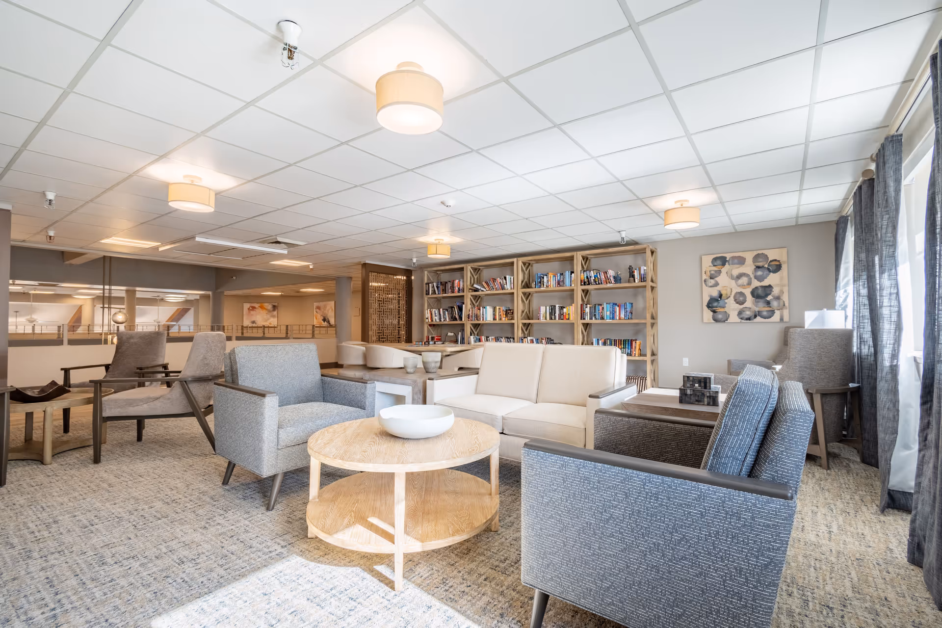 A bright and spacious living room area in a senior living facility with multiple comfortable armchairs and sofas arranged around a round wooden coffee table. There is a large bookshelf filled with books against the back wall, modern ceiling lights, and large windows with sheer curtains allowing natural light to fill the room. The decor includes neutral tones and a piece of abstract wall art.