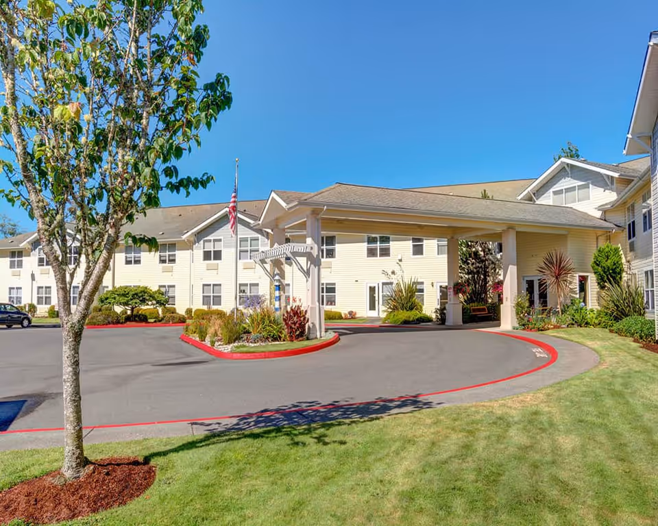 Front entrance and porte-cochère of a multi-story beige senior living building with a circular drive, landscaping, and an American flag.