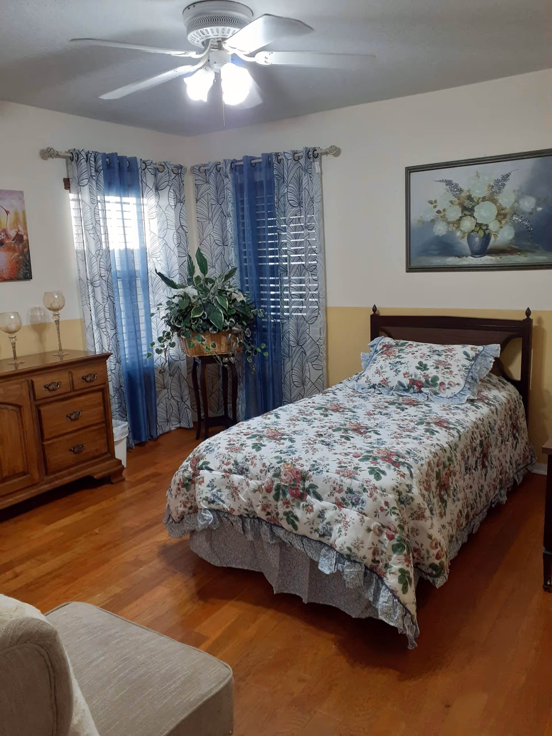 A cozy bedroom with a single bed covered in a floral bedspread. There is a wooden dresser with three glass candle holders on top, a potted plant on a stand near the window, and blue and white patterned curtains. A ceiling fan with lights is mounted on the ceiling, and a framed floral painting hangs on the wall above the bed.