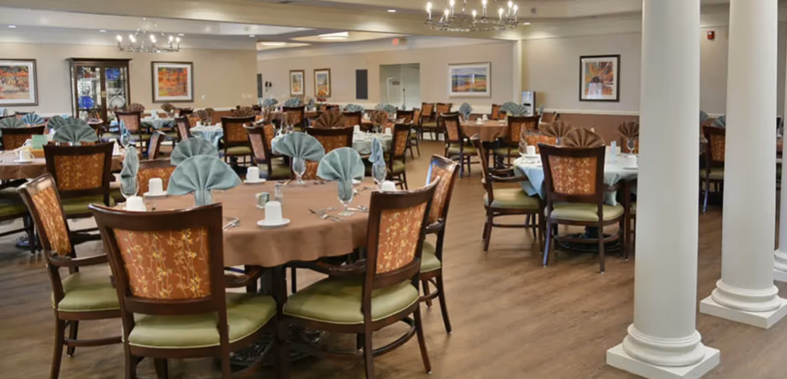 A spacious dining room with multiple round tables covered with brown tablecloths and set with folded napkins, cups, and silverware. The chairs have wooden frames with patterned backs and green cushions. The room has wooden flooring, white columns, and framed artwork on the walls.