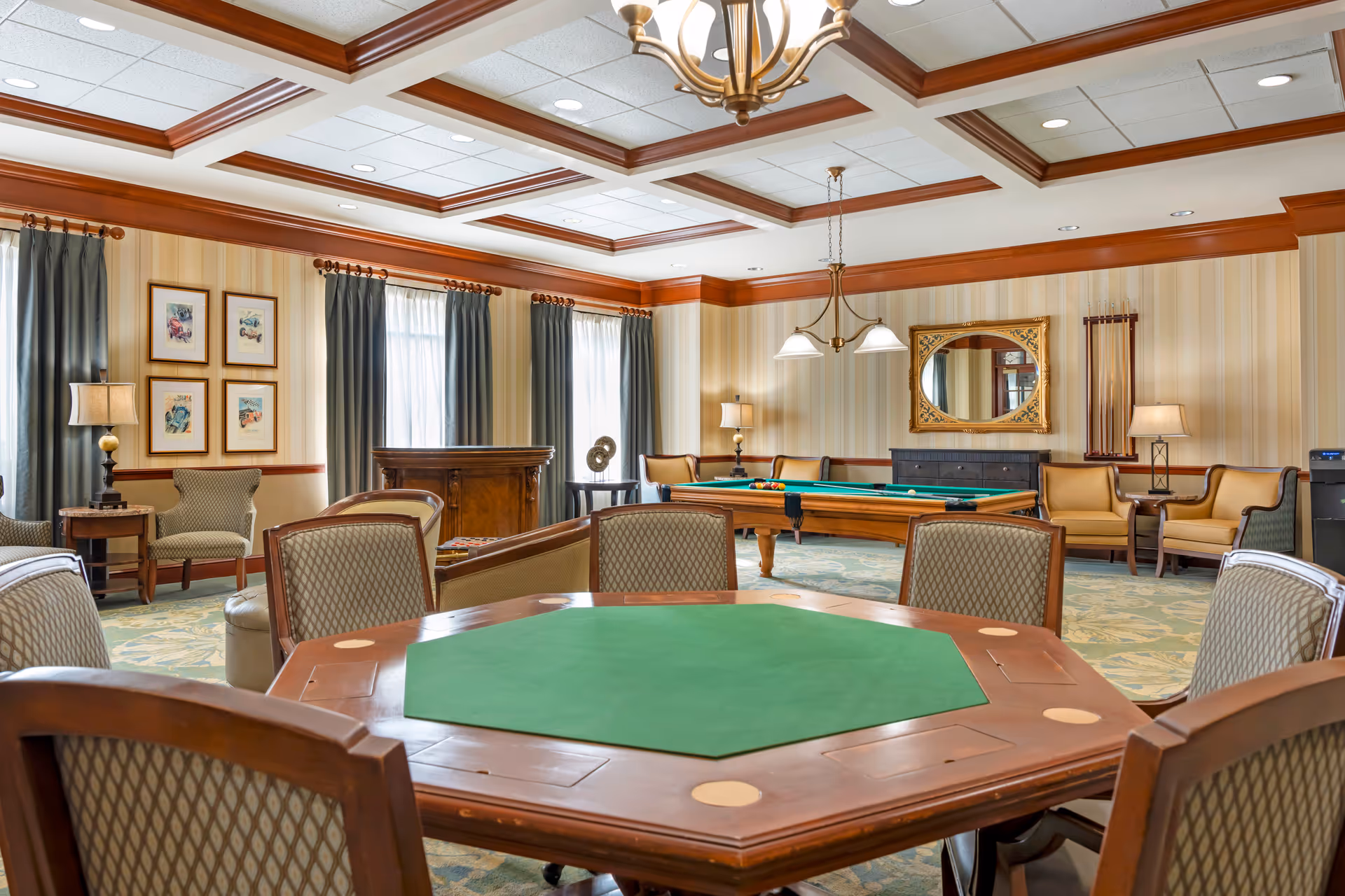 A spacious, well-lit game room with a green felt poker table surrounded by cushioned chairs in the foreground. In the background, there is a pool table with billiard balls arranged, several armchairs and sofas, a large ornate mirror on the wall, framed artwork, and windows with curtains allowing natural light to enter. The ceiling features recessed lighting and wooden beams.