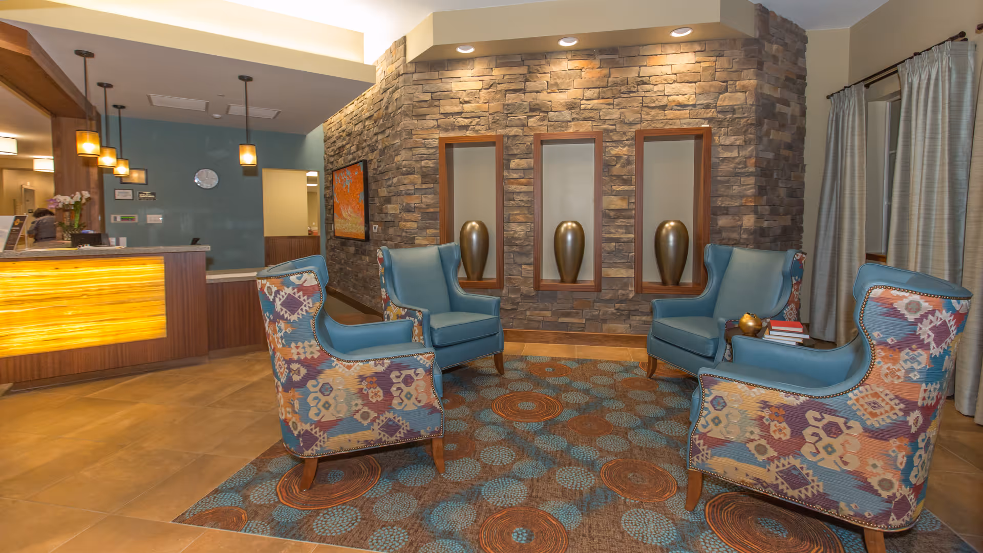 A cozy seating area in a senior living facility lobby with four armchairs arranged around a patterned rug. The chairs have a mix of solid blue and colorful patterned upholstery. Behind the seating area is a stone accent wall with three recessed niches, each holding a decorative vase. To the left is a reception desk with warm lighting and pendant lights hanging from the ceiling. Curtains cover windows on the right side of the room.