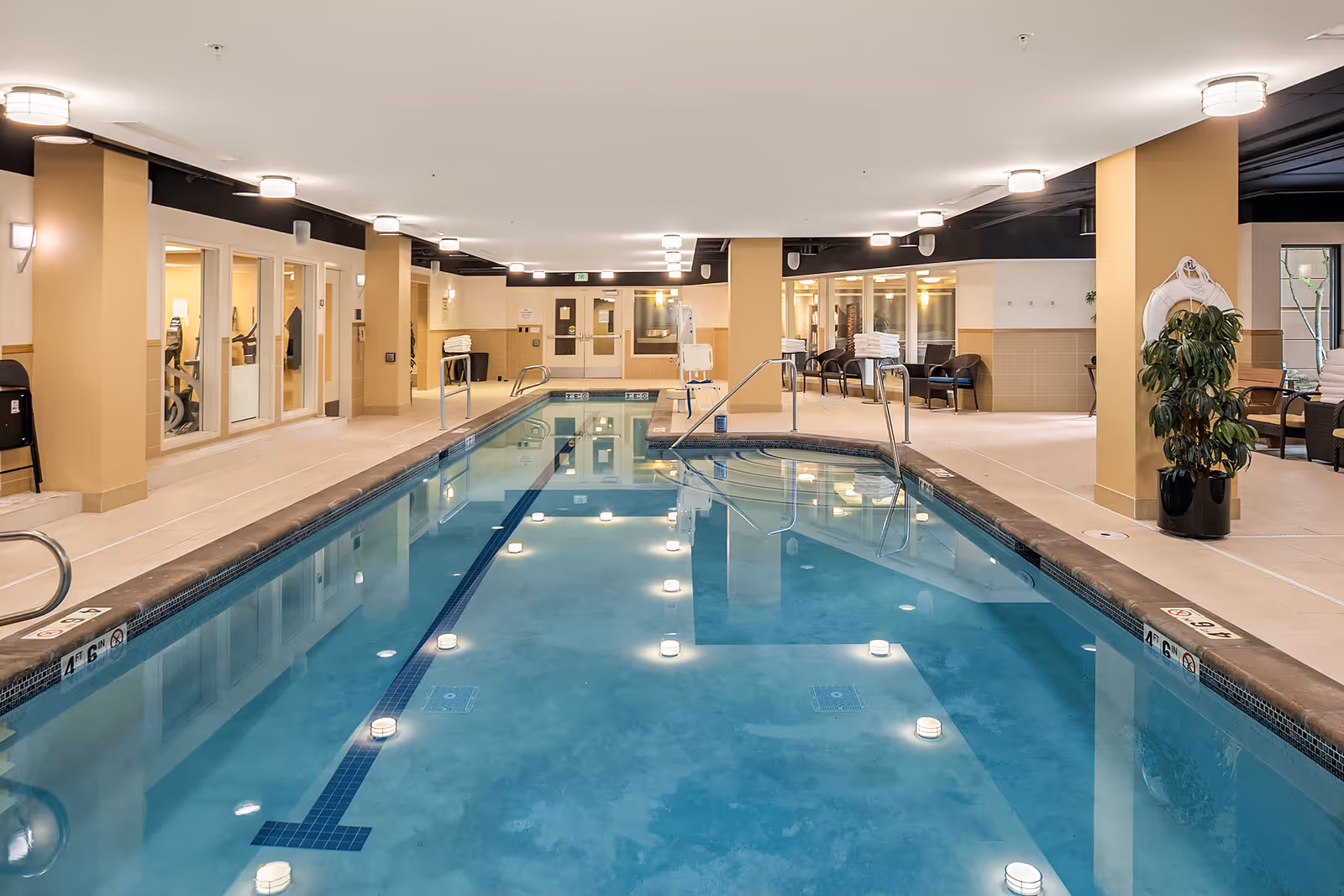 Indoor swimming pool area with clear blue water, surrounded by beige tiled flooring and walls. There are handrails for pool entry, seating areas with chairs and tables, potted plants, and large windows showing an adjacent room with exercise equipment. The ceiling has multiple round light fixtures providing bright illumination.