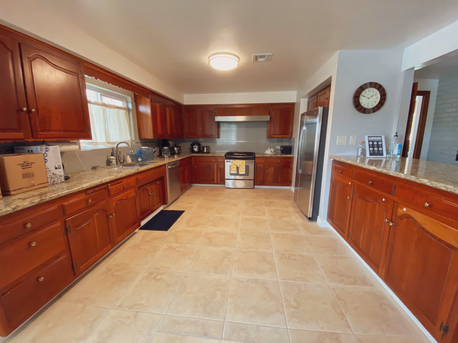 Spacious kitchen with wooden cabinets, granite countertops, tiled floor, stainless steel refrigerator and stove, a clock on the wall, and various kitchen appliances on the counters.