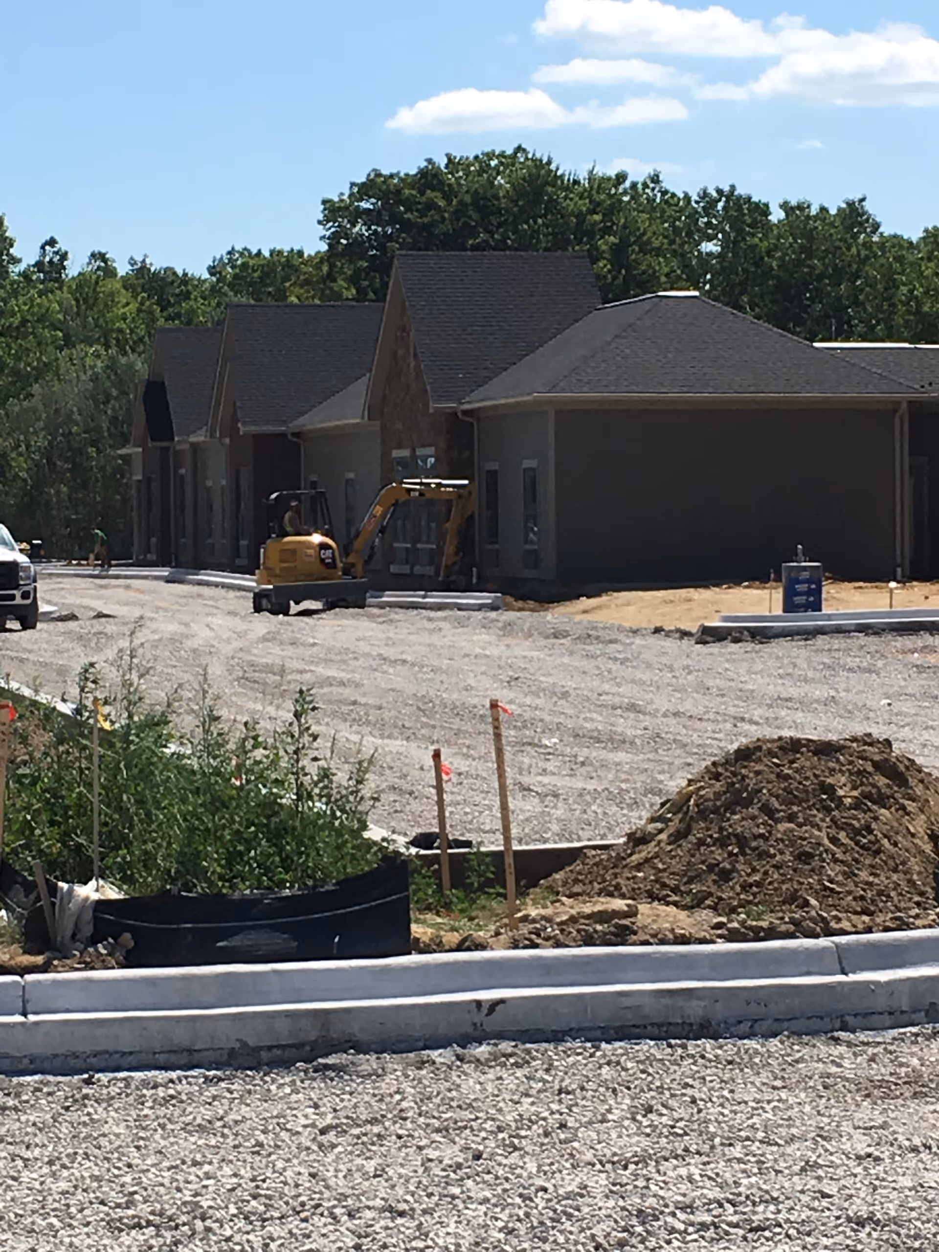 Construction site of a senior living facility with several single-story buildings in progress, a small excavator, gravel road, and some construction materials and dirt piles under a partly cloudy sky.