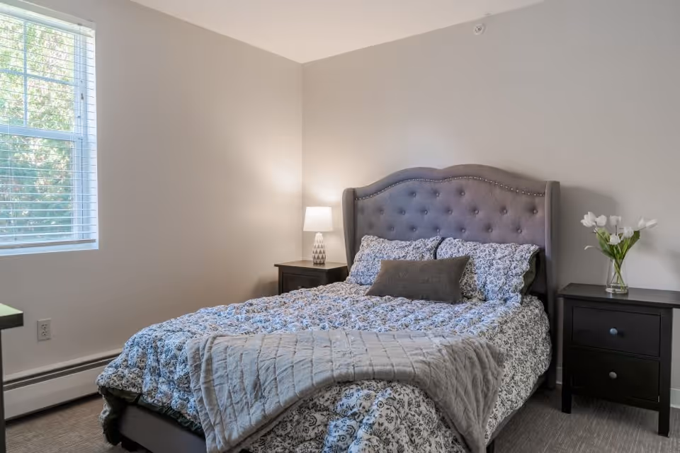 A cozy bedroom with a tufted gray upholstered headboard, a bed with floral patterned bedding and a gray throw blanket. There are two black nightstands on either side of the bed, one with a white table lamp and the other with a vase of white tulips. A window with white blinds allows natural light into the room.