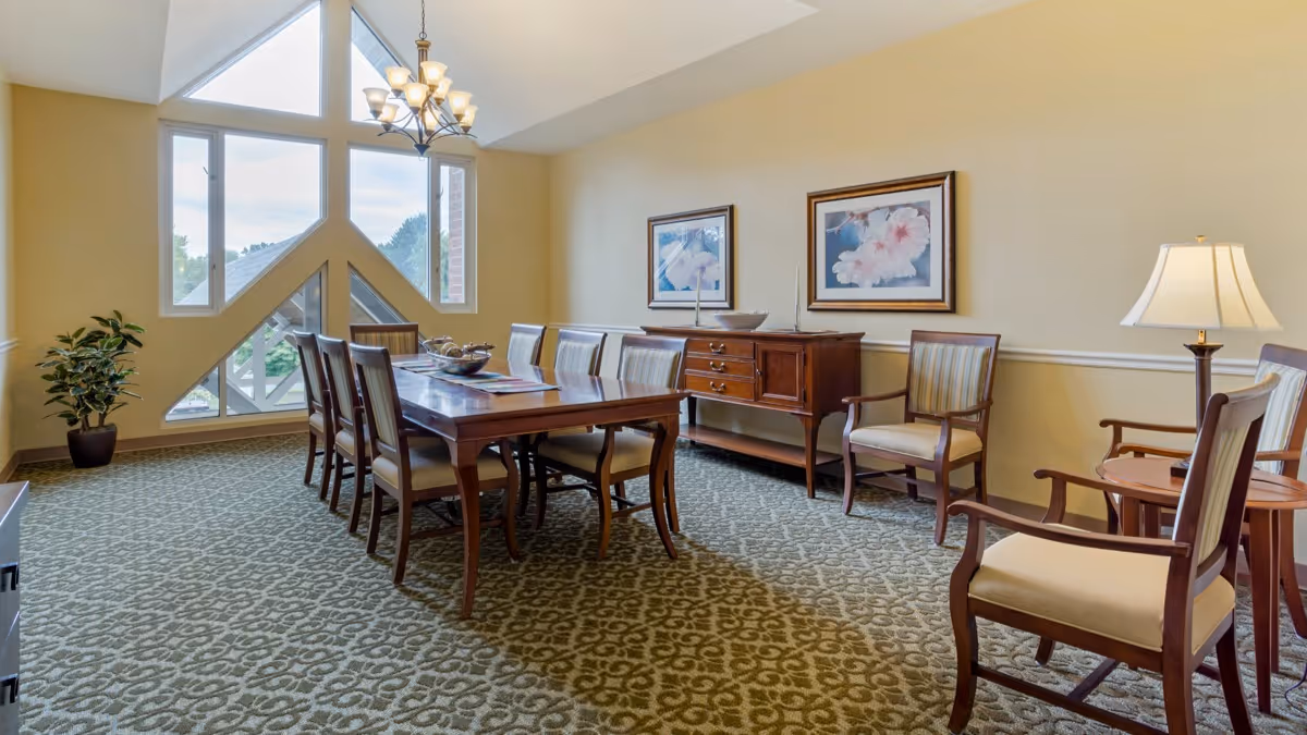 A spacious dining room with a large wooden dining table surrounded by eight chairs. The room features a patterned carpet, a sideboard with decorative items, two framed floral paintings on the wall, a floor lamp, and a large window with triangular panes letting in natural light. A potted plant is placed in the corner.
