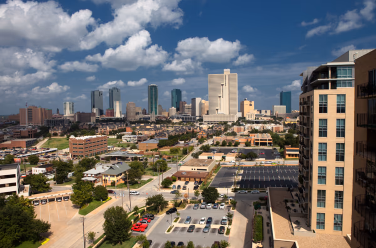 A cityscape view showing a mix of mid-rise and high-rise buildings under a partly cloudy blue sky. In the foreground, there are parking lots with cars, streets, and some trees. On the right side, a tall beige building with many windows is visible.