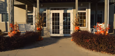Entrance to a building with double glass doors, flanked by windows and decorated with fall-themed arrangements including pumpkins and dried cornstalks. There are bushes and a concrete pathway leading to the doors.