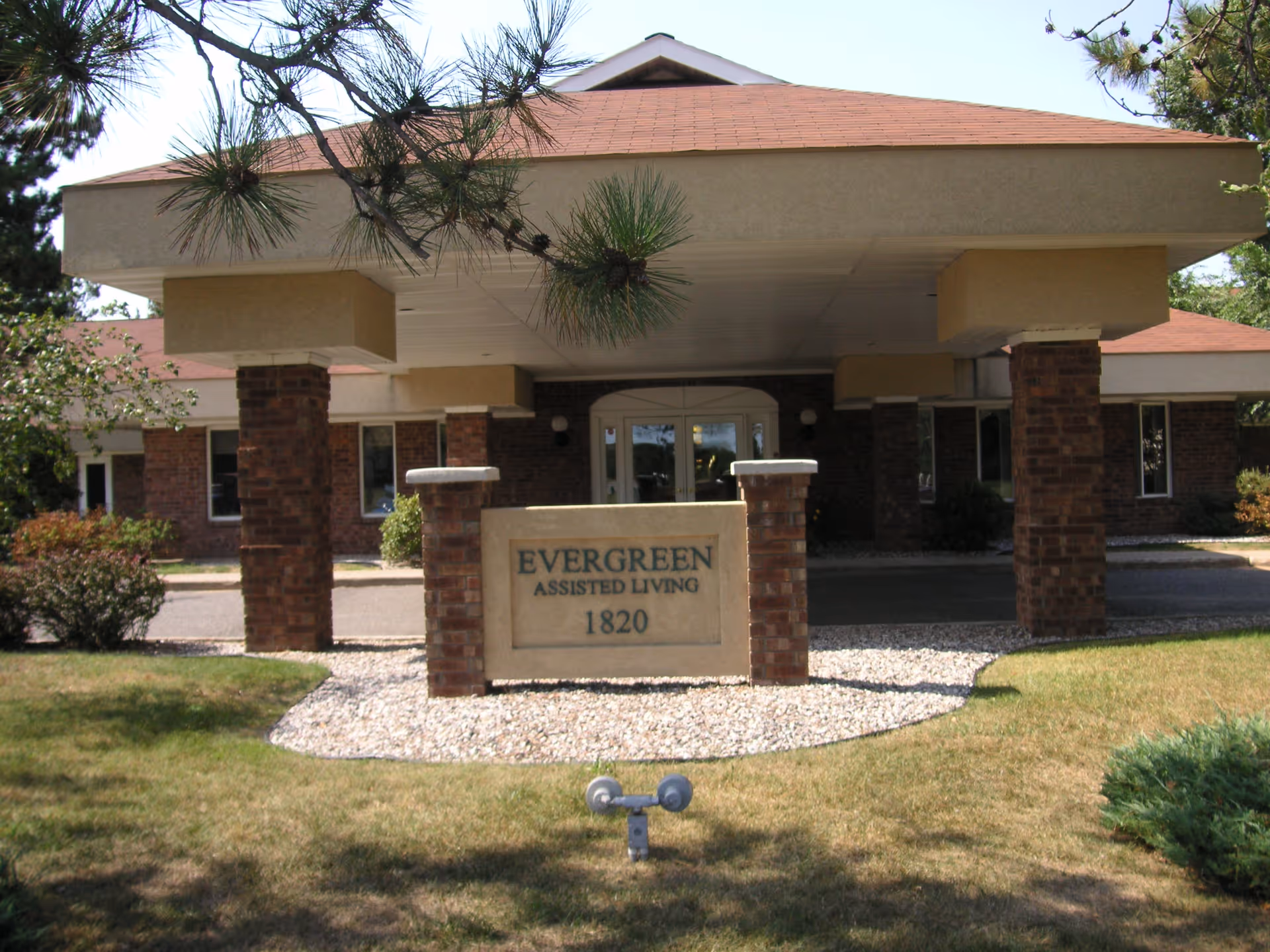 Front entrance of Evergreen Assisted Living facility with a covered drop-off area supported by brick pillars and a sign displaying the facility name and address number 1820. The building has a red roof and is surrounded by grass and some trees.