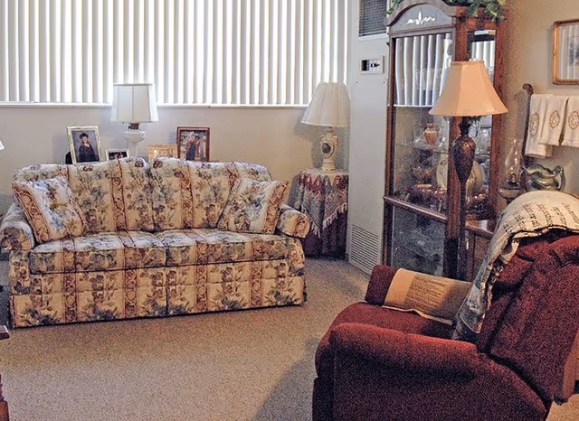 Cozy living room with a floral-patterned sofa, a burgundy recliner, lamps, and a glass display cabinet in front of vertical blinds.