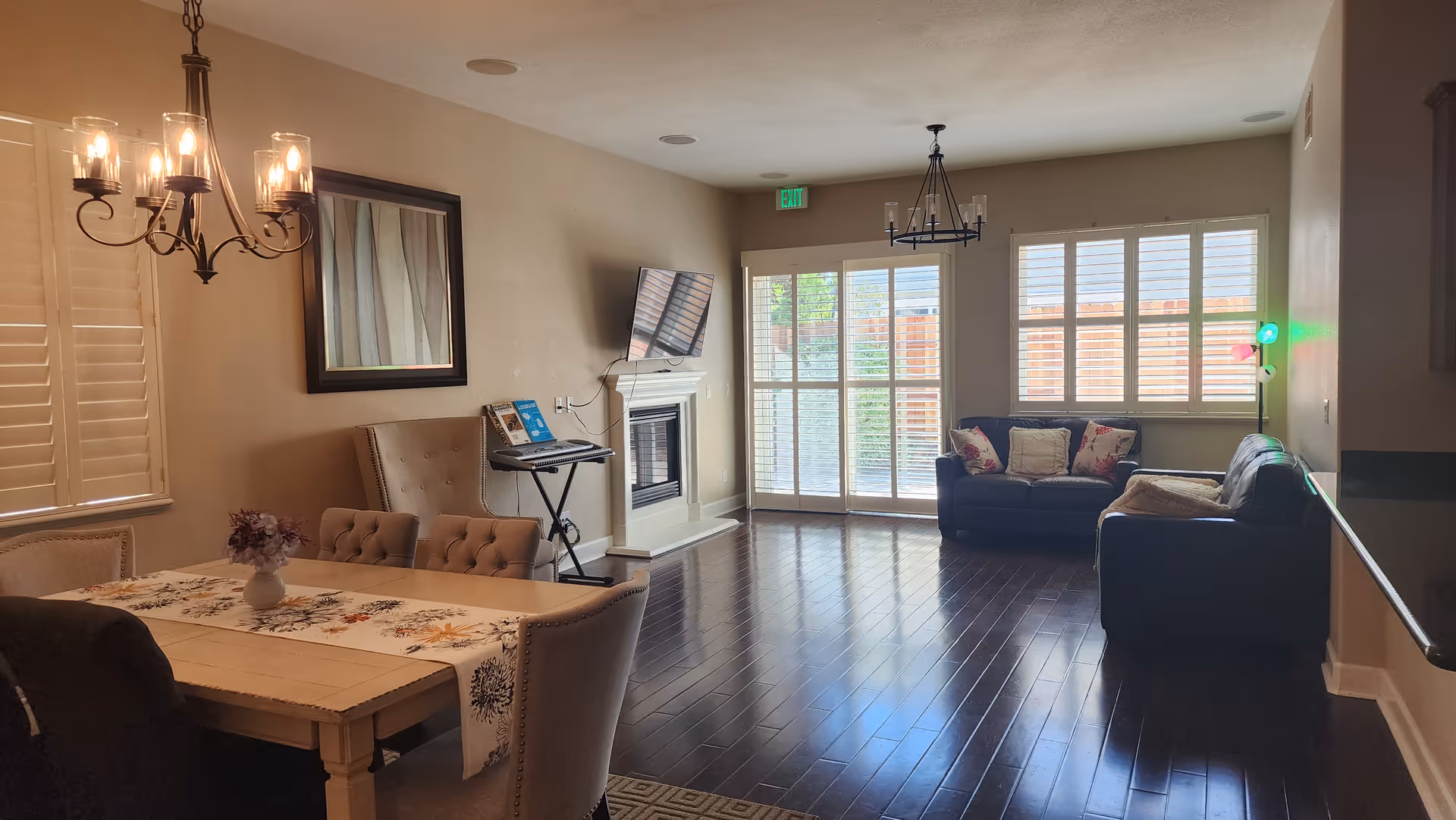 A cozy interior room featuring a dining area with a wooden table, upholstered chairs, and a floral table runner. A chandelier hangs above the table. The room extends into a living area with two dark sofas, decorative pillows, a floor lamp with colored lights, and large windows with white shutters letting in natural light. A wall-mounted TV is above a white fireplace, and there is a keyboard stand next to it. The floor is dark hardwood, and there is an exit sign above a glass door leading outside.