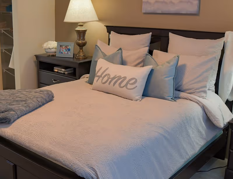 A neatly made bed with a light-colored quilt and multiple pillows, including a decorative pillow with the word 'Home' on it. Next to the bed is a dark wooden nightstand with a lamp, a framed photo, and a small bowl of white flowers. A folded gray blanket is placed at the foot of the bed.