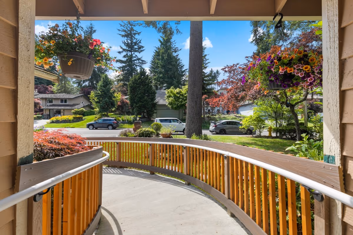 View from a covered porch or entrance ramp with wooden railings and hanging flower pots, overlooking a suburban street with trees, parked cars, and houses under a blue sky with some clouds.