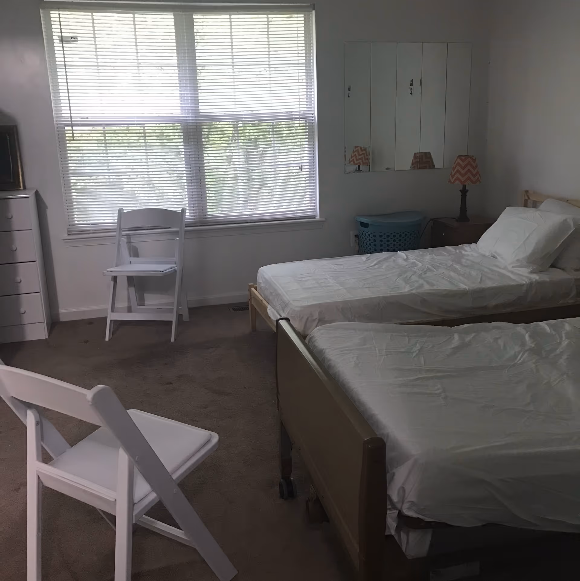 A simple bedroom with two single beds covered in white sheets, two white folding chairs, a window with blinds, a dresser, a nightstand with a lamp, and a laundry basket in the corner.