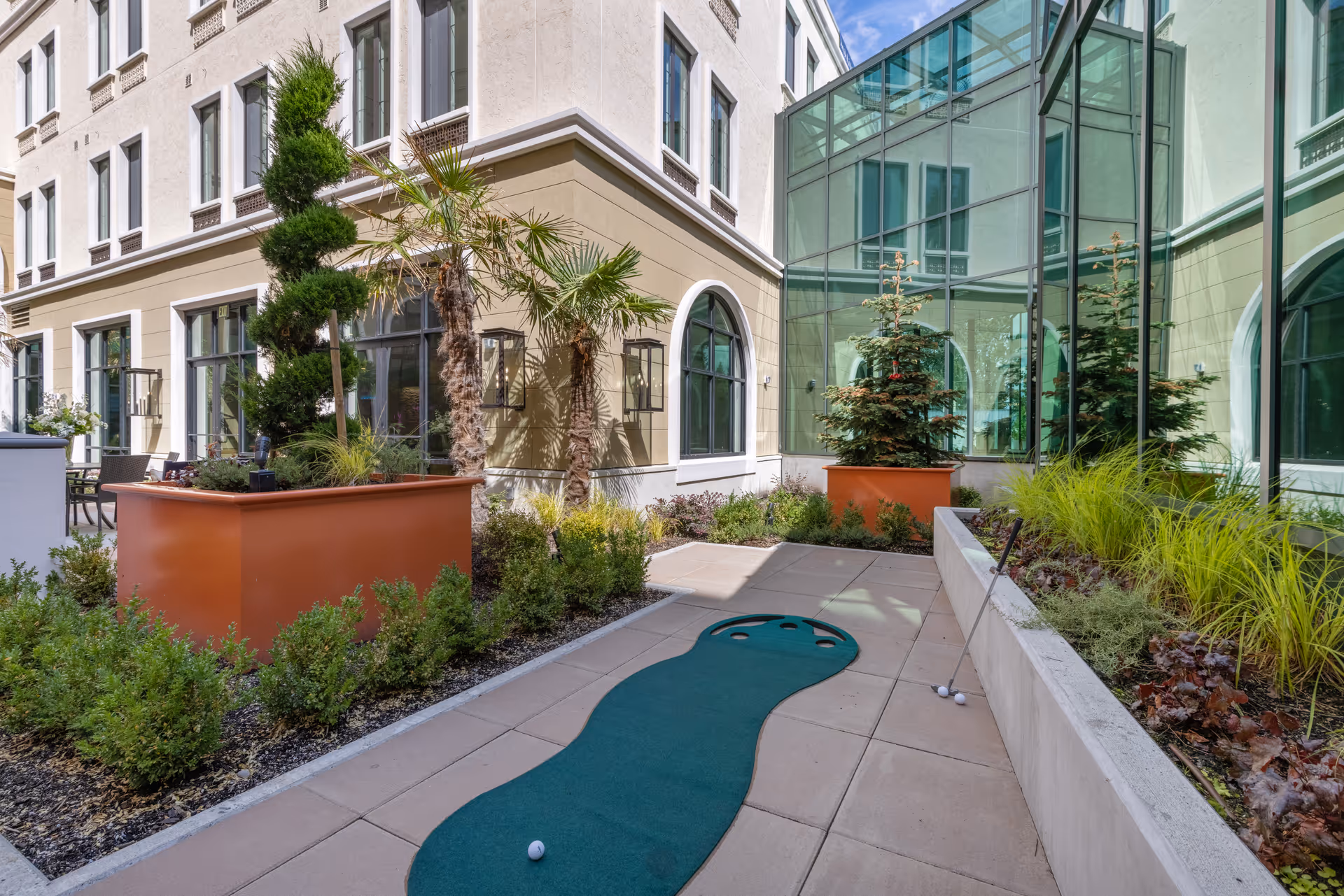 Sunny courtyard with a small putting green, large planters and landscaping beside a multi-story building and glass atrium.