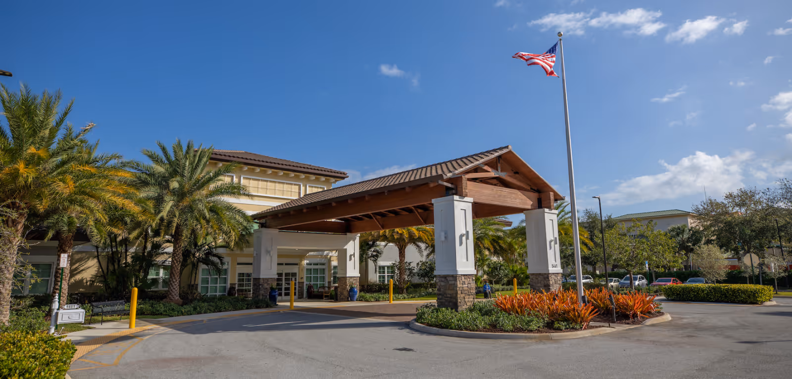 Covered entrance and porte-cochère of a senior living facility with palm trees and an American flag in front.