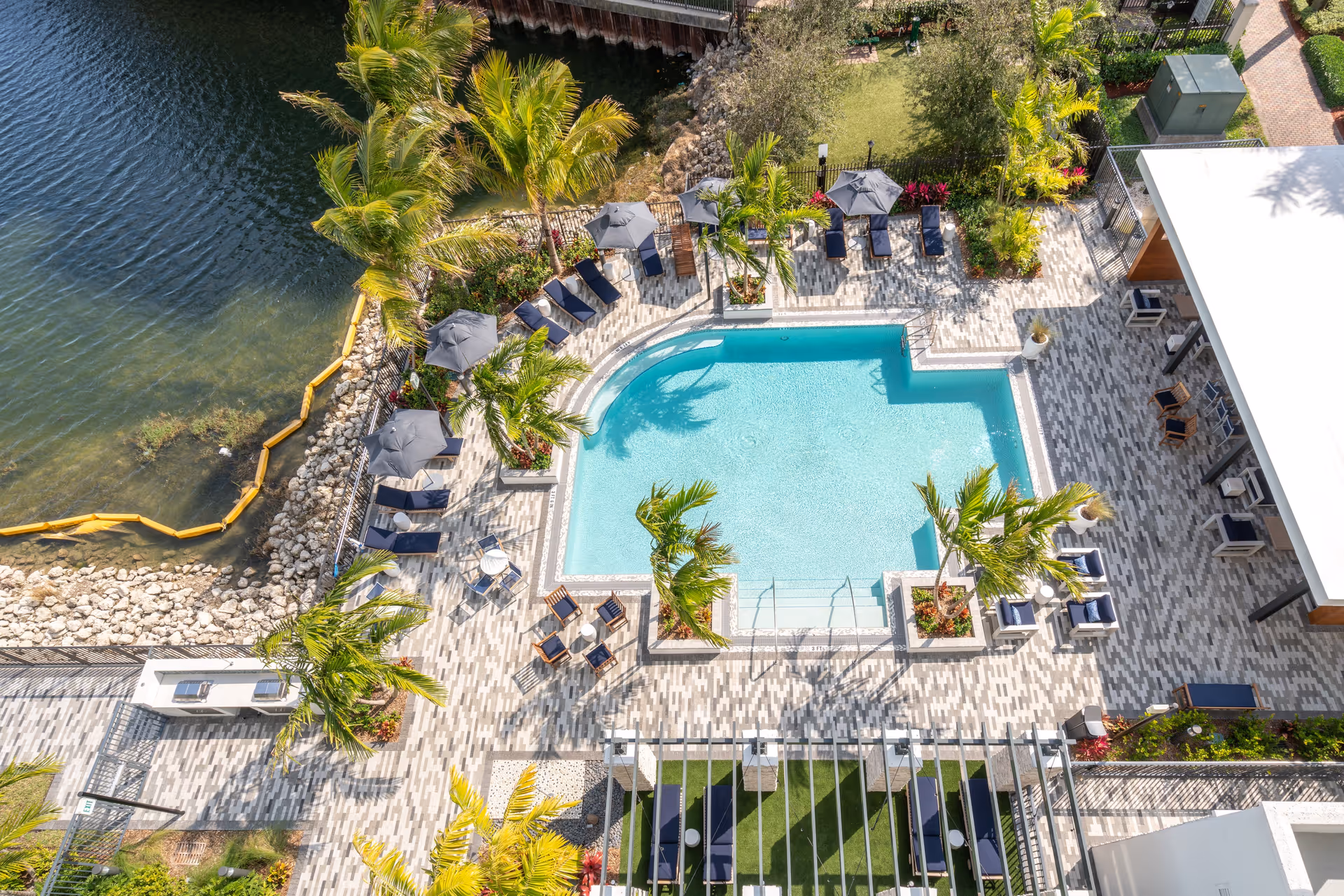 Aerial view of a swimming pool area surrounded by lounge chairs, umbrellas, palm trees, and a tiled patio. The pool is adjacent to a body of water with a rocky shoreline and yellow floating barriers. There is also a covered seating area with chairs and tables on the right side of the image.