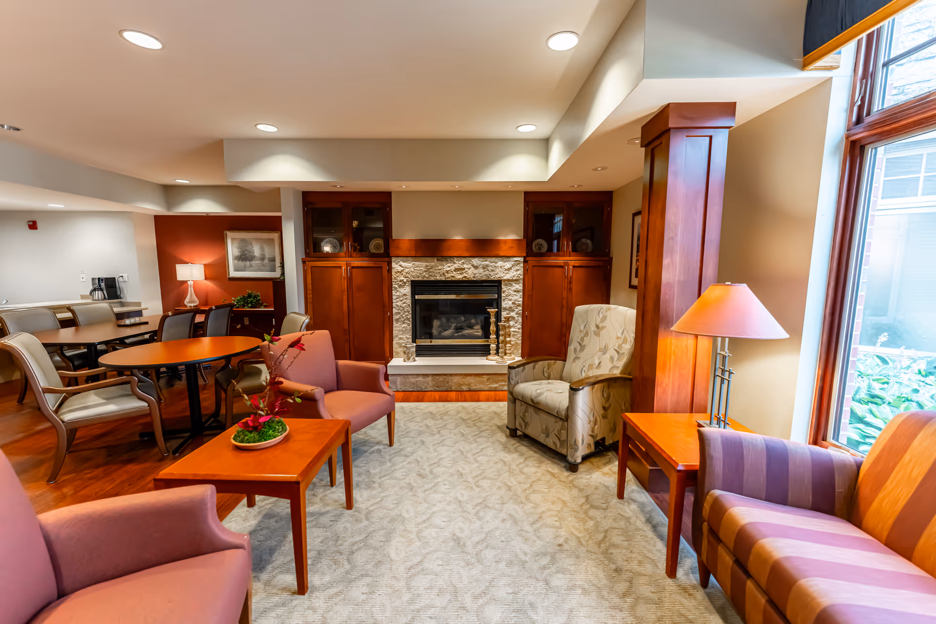 Cozy common living area with upholstered chairs, tables, a fireplace, and a large window.