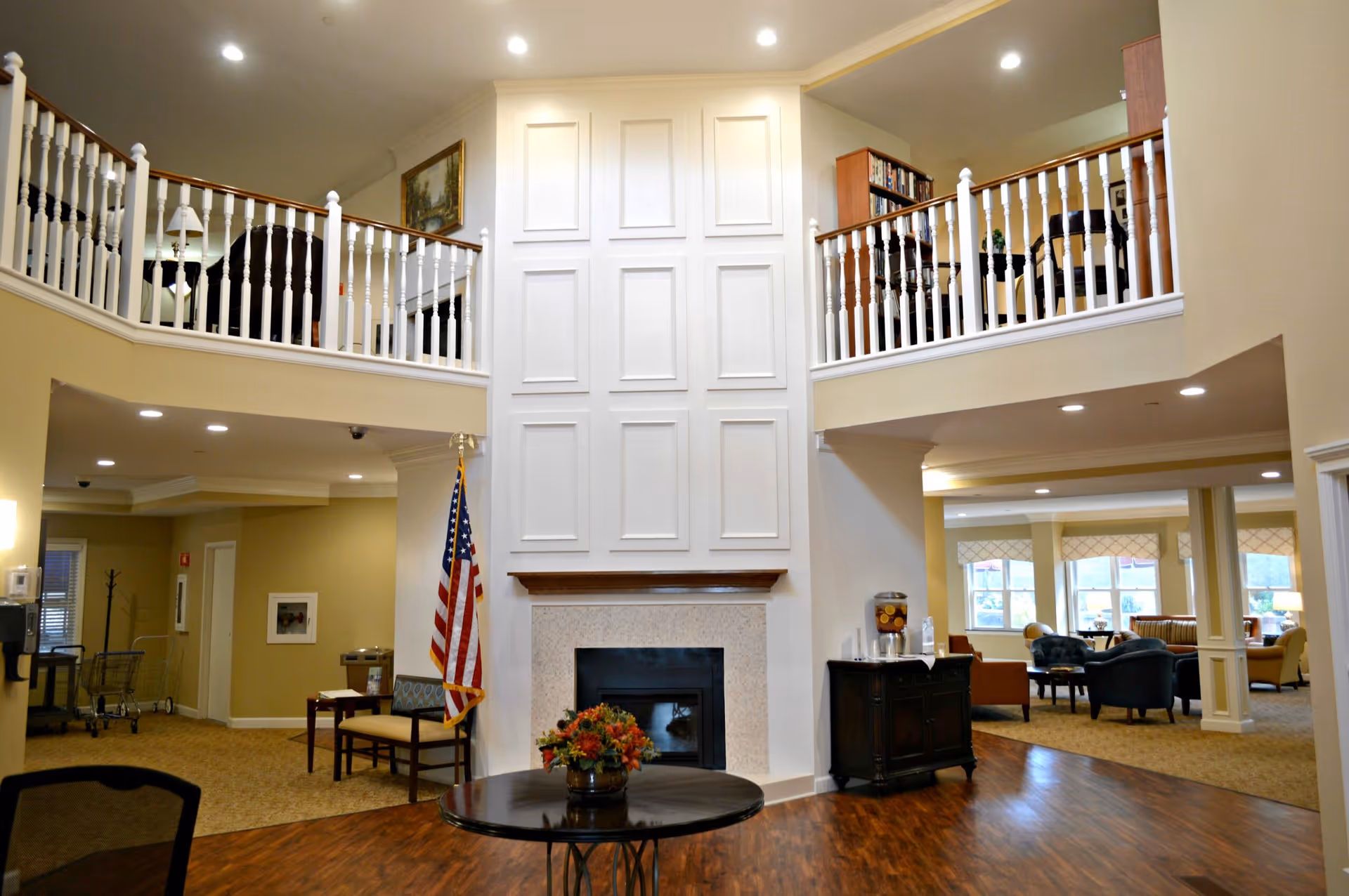 Interior view of a senior living facility lobby area with a central fireplace, a round table with a flower arrangement, an American flag, and seating areas on both the main floor and an upper balcony. The space is well-lit with recessed lighting and has a warm, welcoming atmosphere.