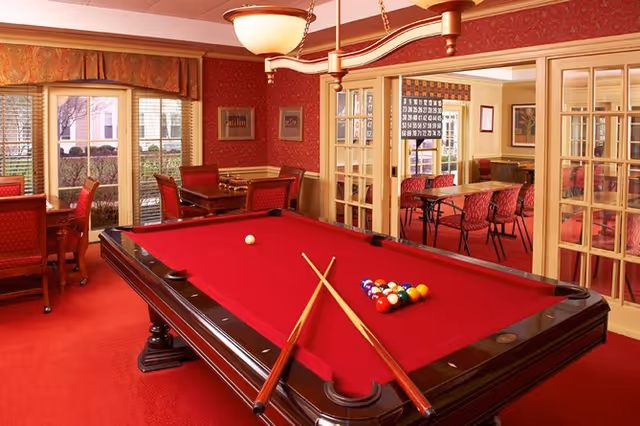 A cozy game room with a red pool table in the center, two pool cues resting on the table, and a rack of billiard balls arranged for a game. The room has red carpet and walls with framed pictures, a large window with a view of greenery outside, and a separate adjoining room visible through glass-paneled double doors containing tables and chairs.