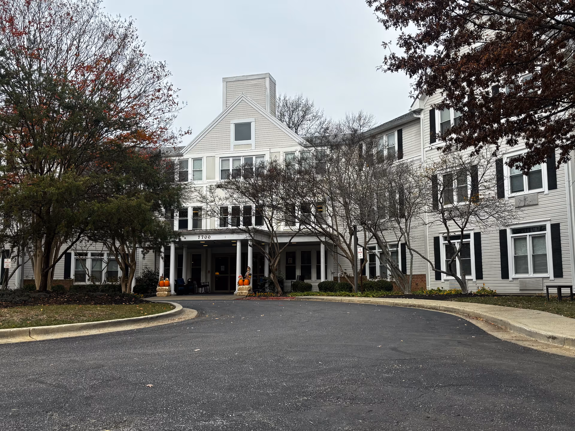Front entrance of a multi-story assisted living building with a circular driveway, trees, and fall decorations.