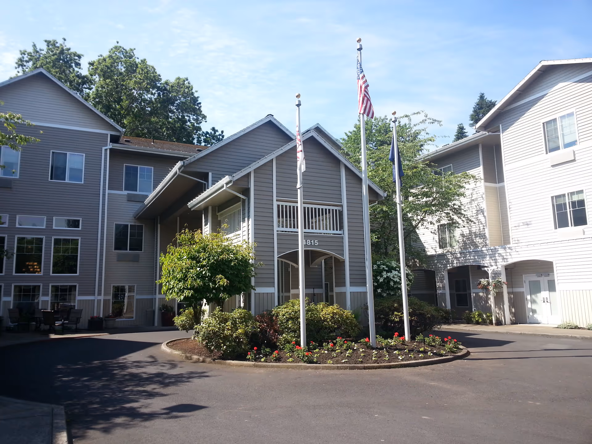 Exterior view of a multi-story assisted living facility building with gray siding, multiple windows, and a covered entrance. There are three flagpoles with flags in front of the building, surrounded by landscaped bushes and flowers. The sky is clear and blue.