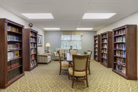 A well-lit library room with multiple wooden bookshelves filled with books along the walls. In the center, there is a wooden table surrounded by four cushioned chairs. At the back of the room, there are two armchairs and a small table with a lamp, positioned near a window with a valance. The carpet has a patterned design, and the ceiling has recessed lighting.