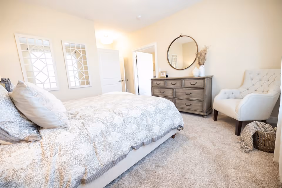 Bright, neutrally decorated bedroom featuring a bed with patterned bedding, a wooden dresser topped by a round mirror, and a tufted armchair.