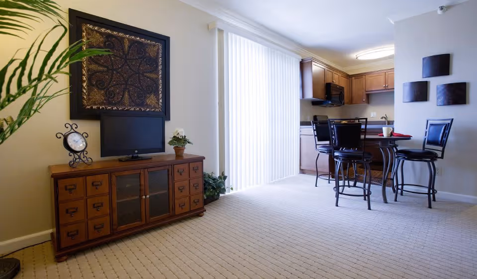 A bright interior space featuring a wooden cabinet with a small TV, decorative clock, and potted plant on top. To the right, there is a dining area with a round table and four high chairs. The kitchen with wooden cabinets and appliances is visible in the background. Vertical blinds cover a large window or sliding door, allowing natural light to fill the room.