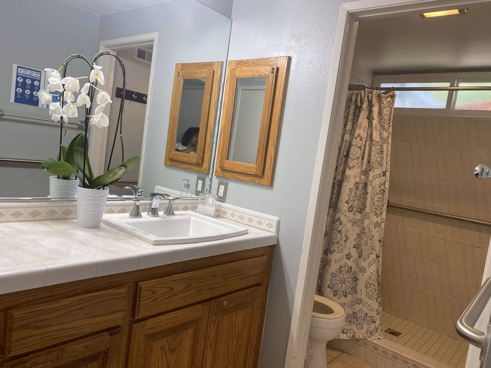 Bathroom with a wooden vanity featuring a white sink and a potted orchid plant on the countertop. There is a large mirror above the sink and two wooden framed medicine cabinets on the adjacent wall. To the right, there is a shower area with a floral-patterned shower curtain and a toilet partially visible. The walls are light-colored and there are grab bars installed near the shower and toilet.