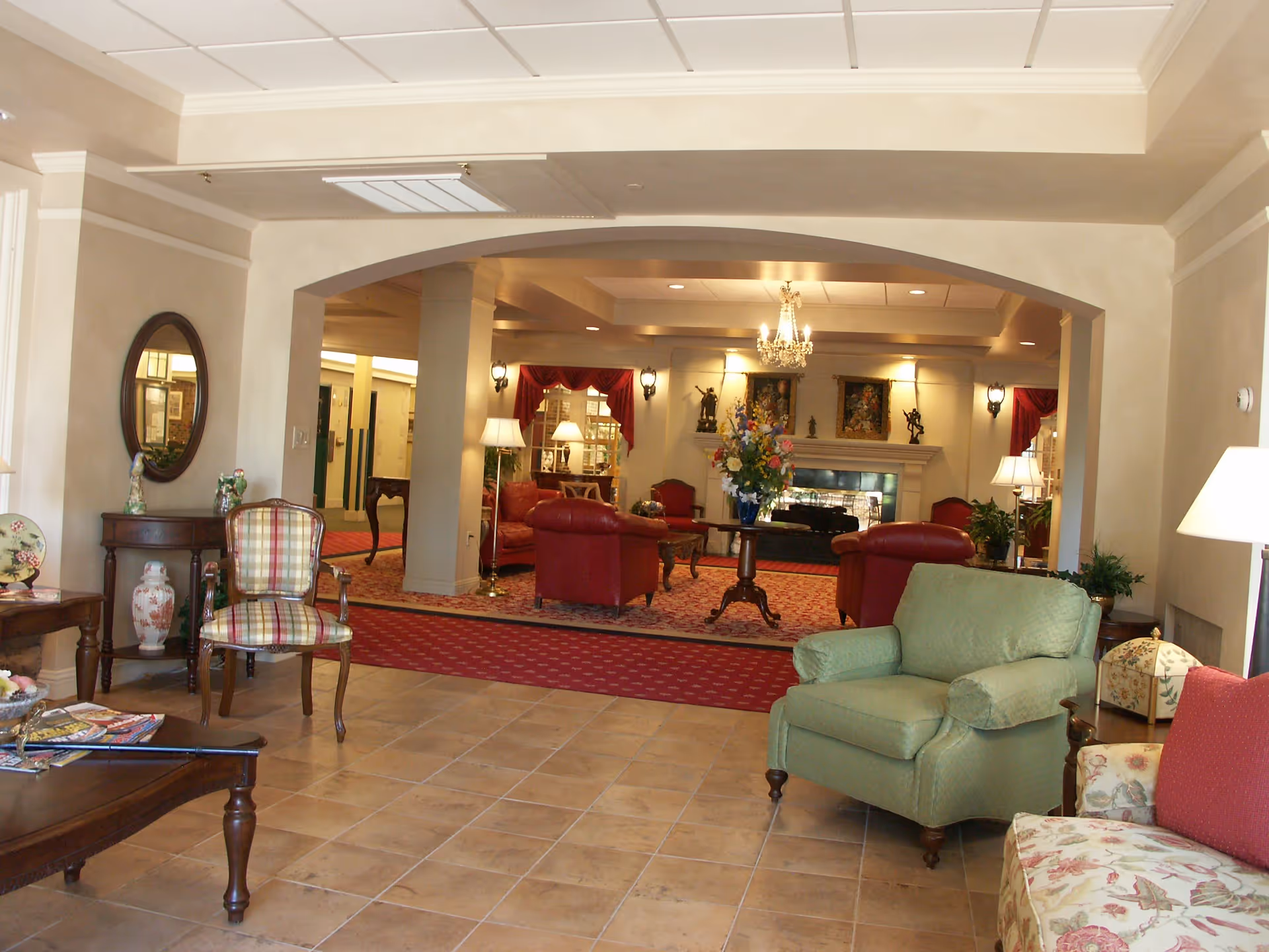 A spacious and warmly decorated living room area in an assisted living facility. The room features a tiled floor in the foreground with a green armchair and a floral-patterned sofa. In the background, there is a carpeted seating area with red leather chairs, a round wooden table with a flower arrangement, and a fireplace adorned with artwork and sculptures. The room is softly lit with table lamps and a chandelier, and there is a large archway separating the two areas.