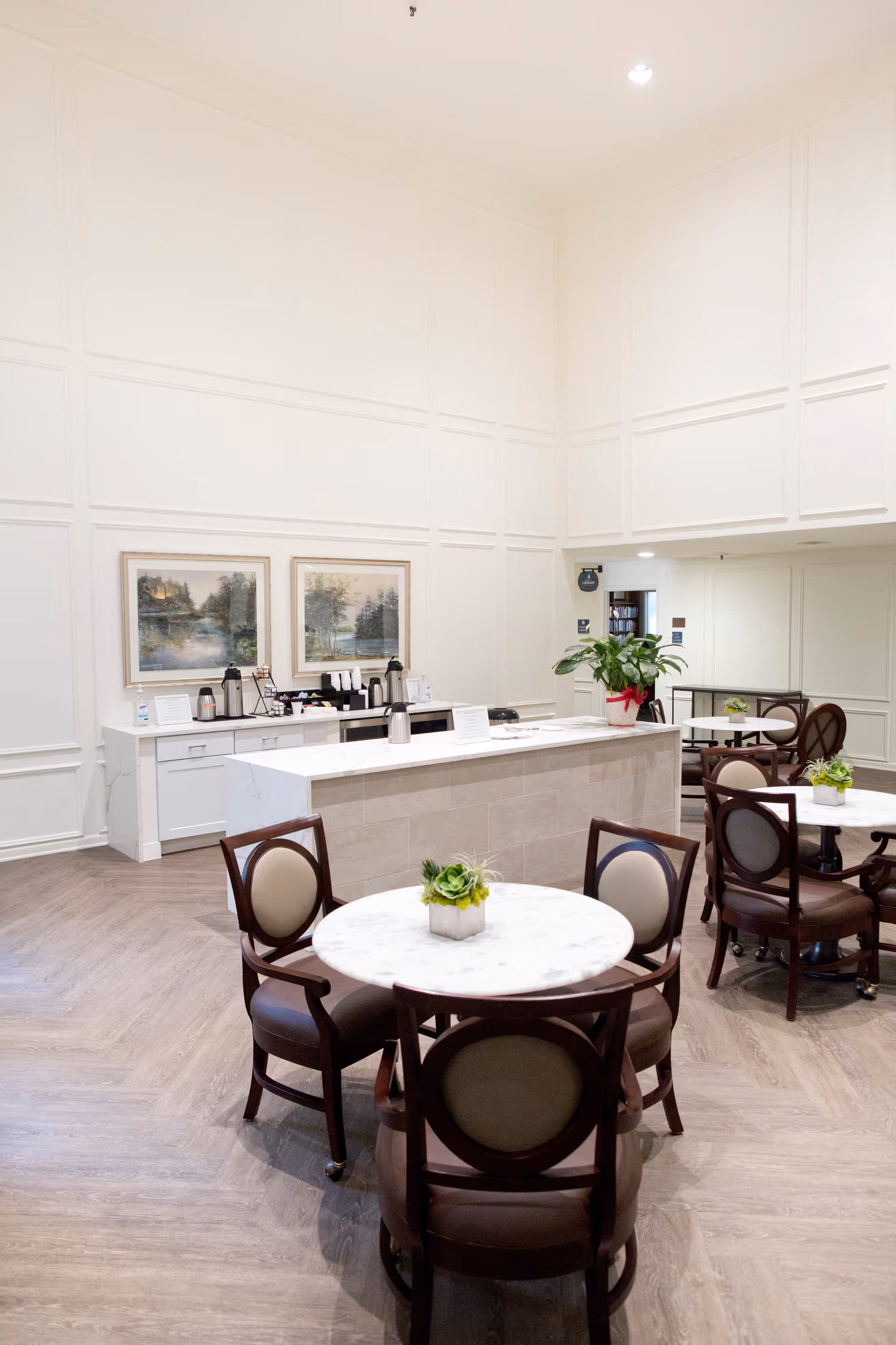 A bright and clean common area with round marble tables and wooden chairs with cushioned seats. A counter with coffee and beverage supplies is against the wall, decorated with two landscape paintings and a potted plant. The room has high ceilings and white paneled walls.