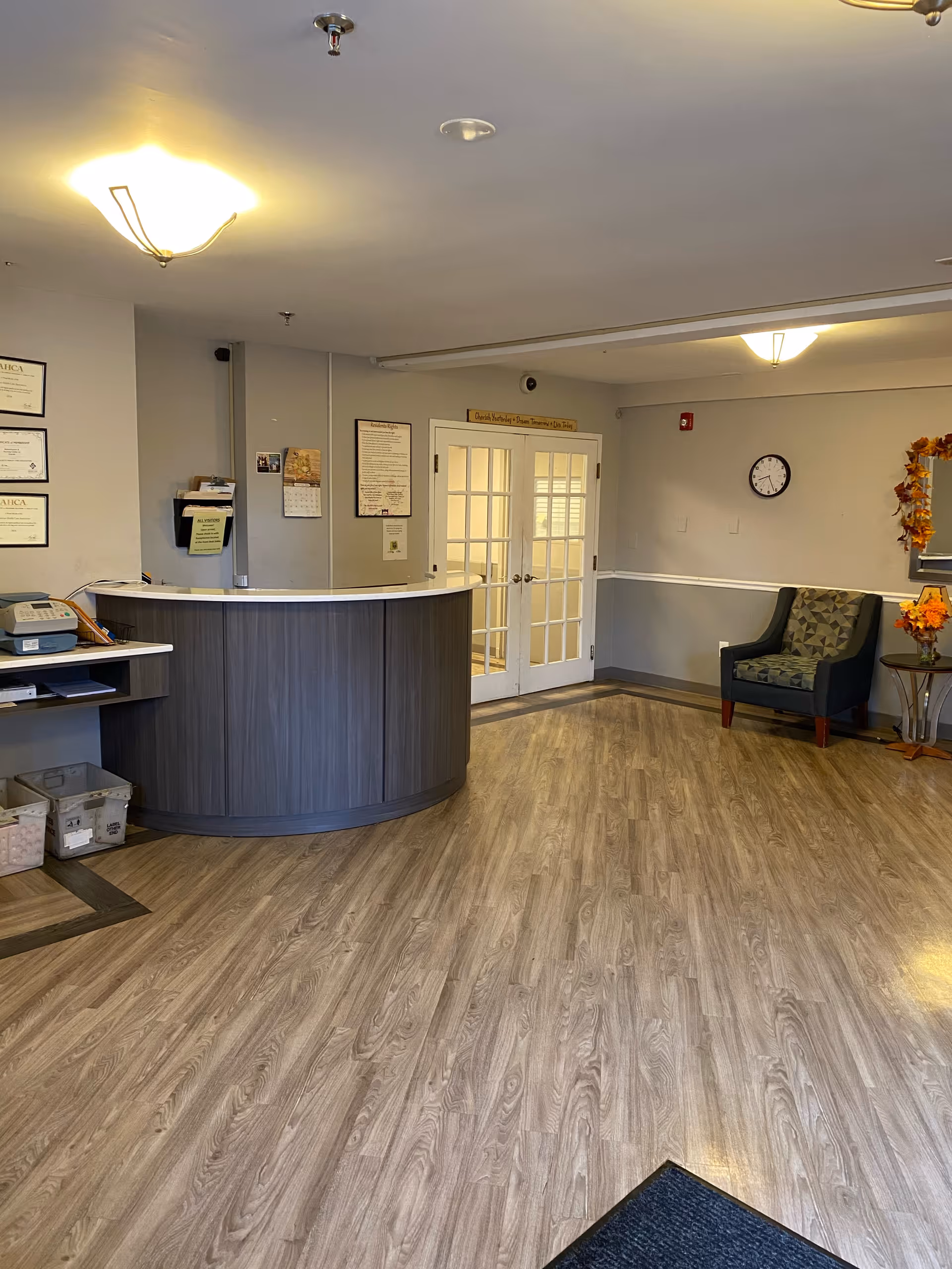 A clean reception lobby with a curved front desk, wood-look flooring, an armchair, clock on the wall and double glass doors.