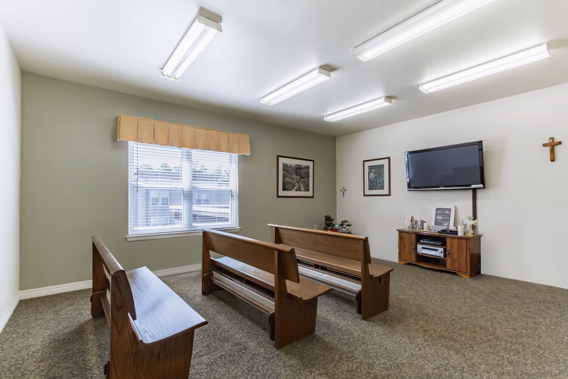 A small room with three wooden pew-style benches facing a wall-mounted flat screen TV. The room has a window with blinds and a valance, two framed pictures on the walls, a wooden cabinet under the TV holding various items, and a cross hanging on the wall. The floor is carpeted and the ceiling has fluorescent lights.