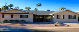Single-story building with a beige exterior and a gray roof, surrounded by palm trees under a clear blue sky, viewed from across the street.