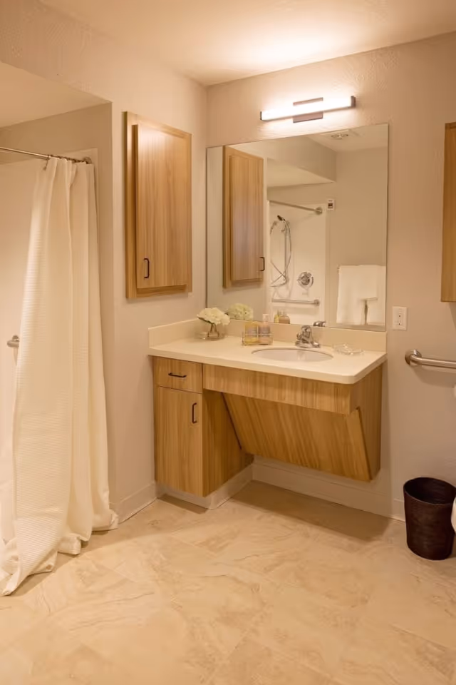 A clean and well-lit bathroom featuring a wooden vanity with a white countertop and sink, a large mirror above the sink, a wall-mounted cabinet, a shower area with a white curtain, and a small black trash bin on the floor.