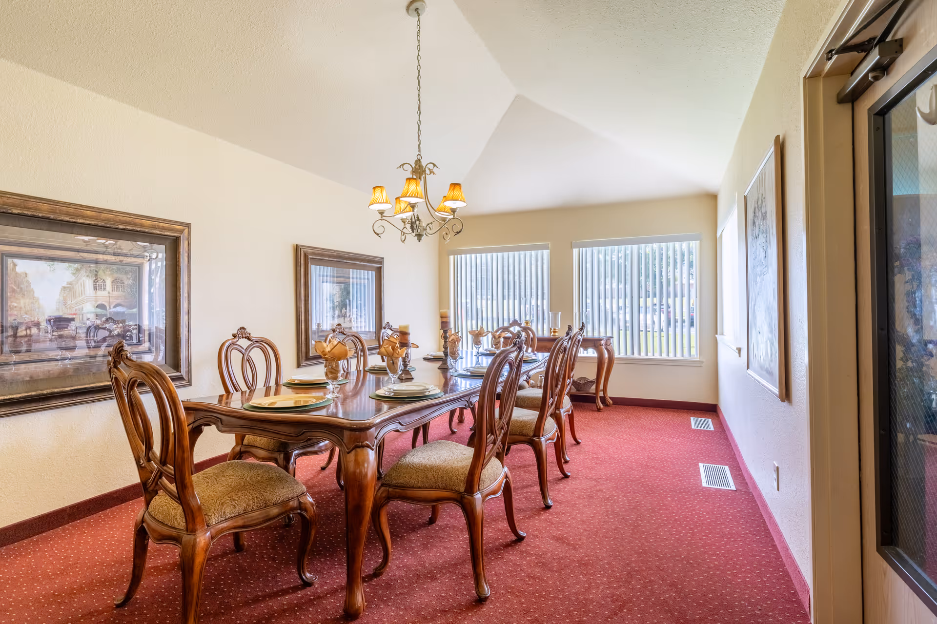 Formal dining room with a long wooden table and chairs under a chandelier, framed artwork on the walls, and windows with vertical blinds.