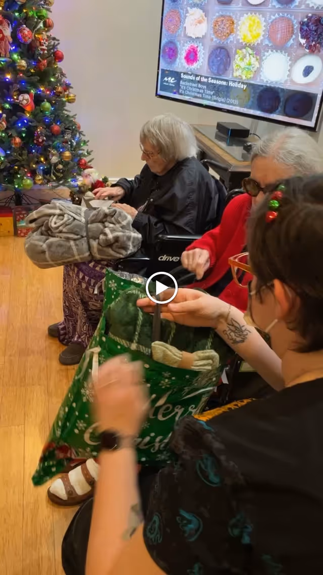 Residents seated by a decorated Christmas tree exchanging gifts in a communal living room.